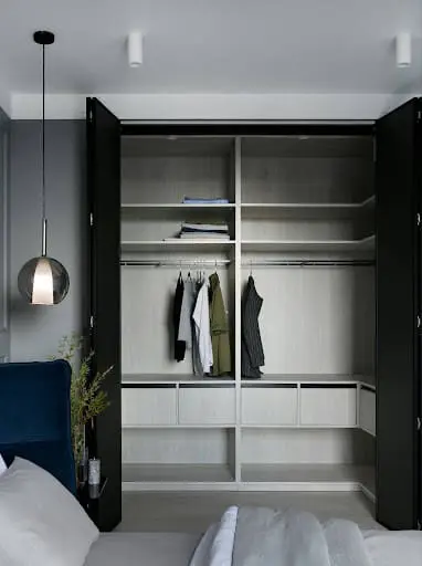 A modern bespoke built-in wardrobe featuring a white laminate interior with multiple shelving levels, a hanging rail with clothes, and integrated lighting, viewed through open dark-colored doors.