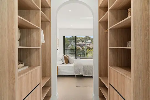 Custom light-timber melamine walk-in wardrobe featuring vertical shelving compartments, hanging rails, and an arched passageway overlooking a bright bedroom with a view