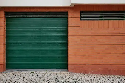 A closed dark green roller garage door on a brick driveway, framed by light-colored wooden wall cladding